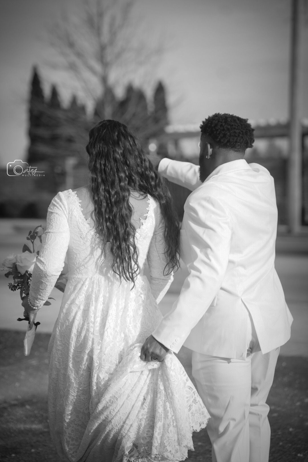 Black and white photo of newlyweds walking together in elegant white wedding attire.