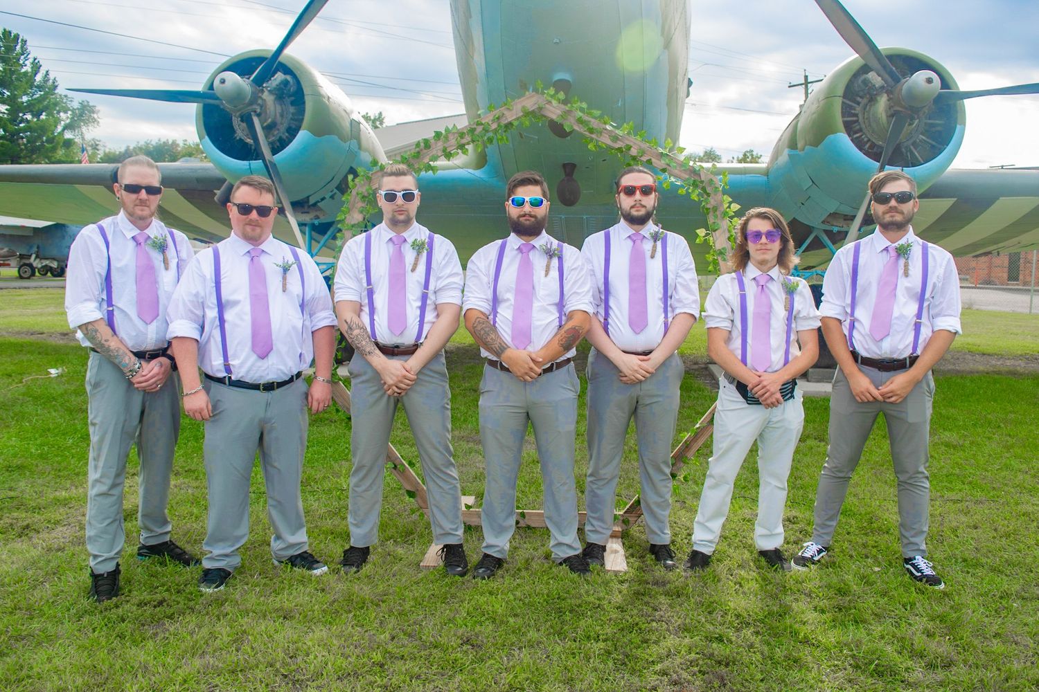 Groomsmen in light gray suits and suspenders line up for photo by vintage airplane.