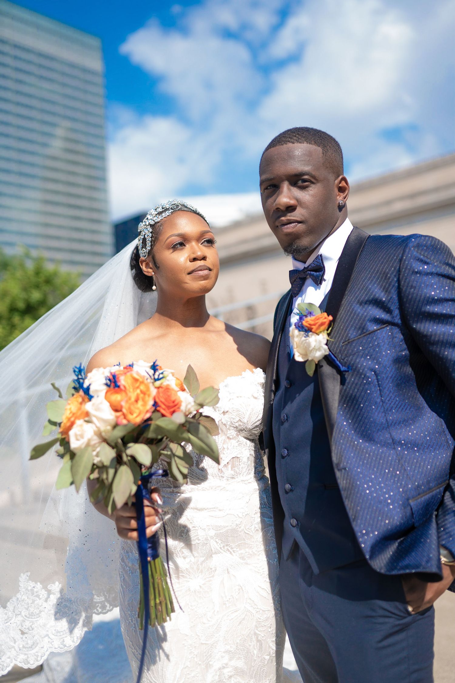 Elegant wedding couple poses outdoors with orange and blue floral bouquet against blue sky.