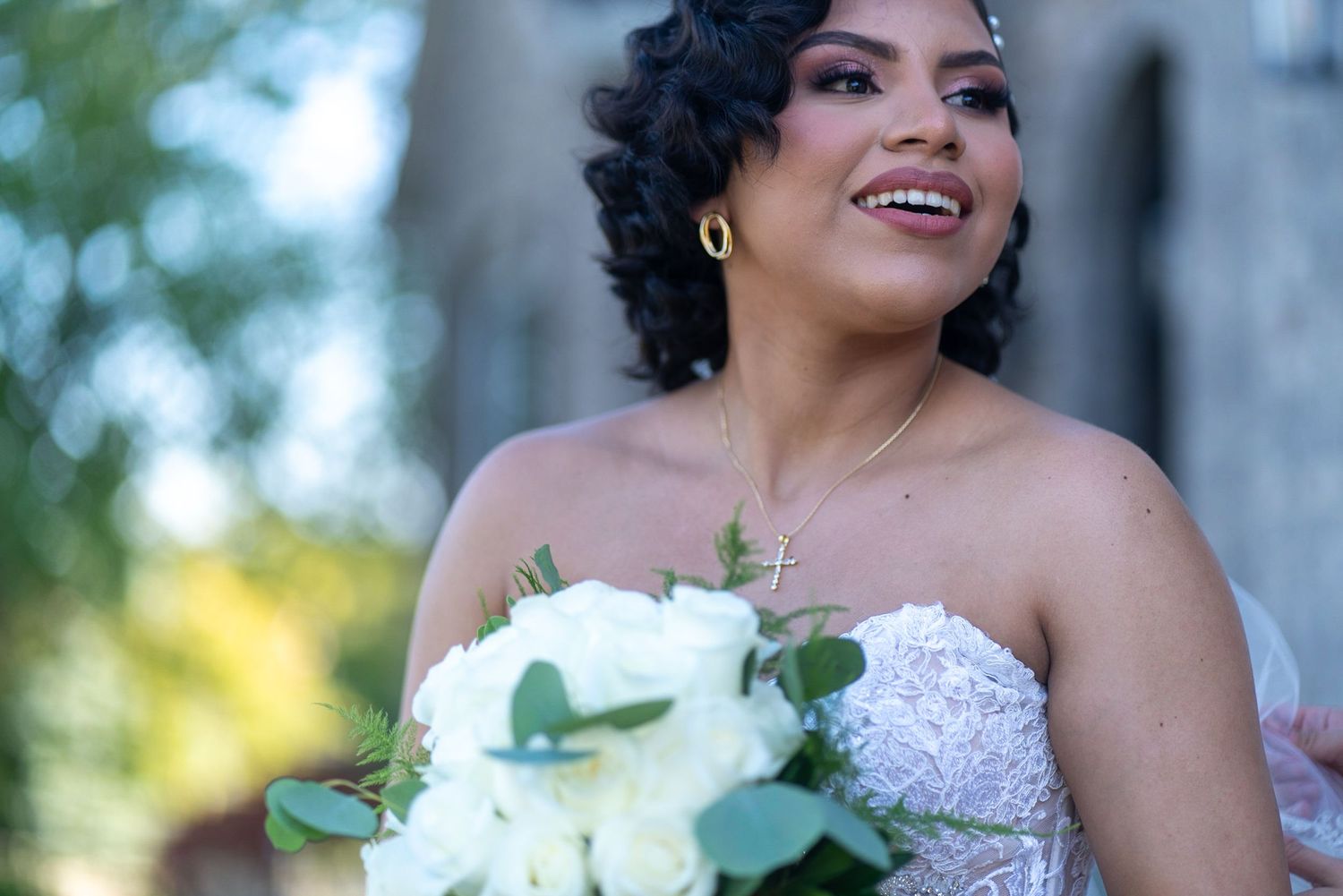 A bride in a white lace gown smiles while holding a bouquet of white flowers.