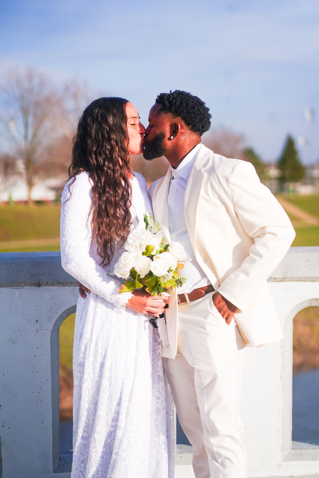 Couple in white attire shares romantic moment by waterfront during wedding ceremony.
