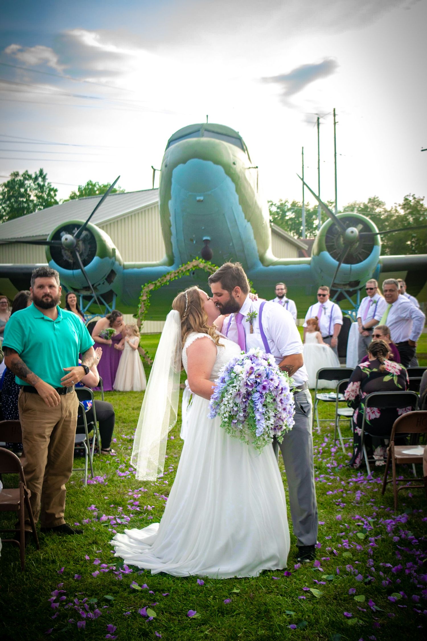 Wedding ceremony taking place in front of a vintage turquoise airplane with purple flowers decorating the scene.
