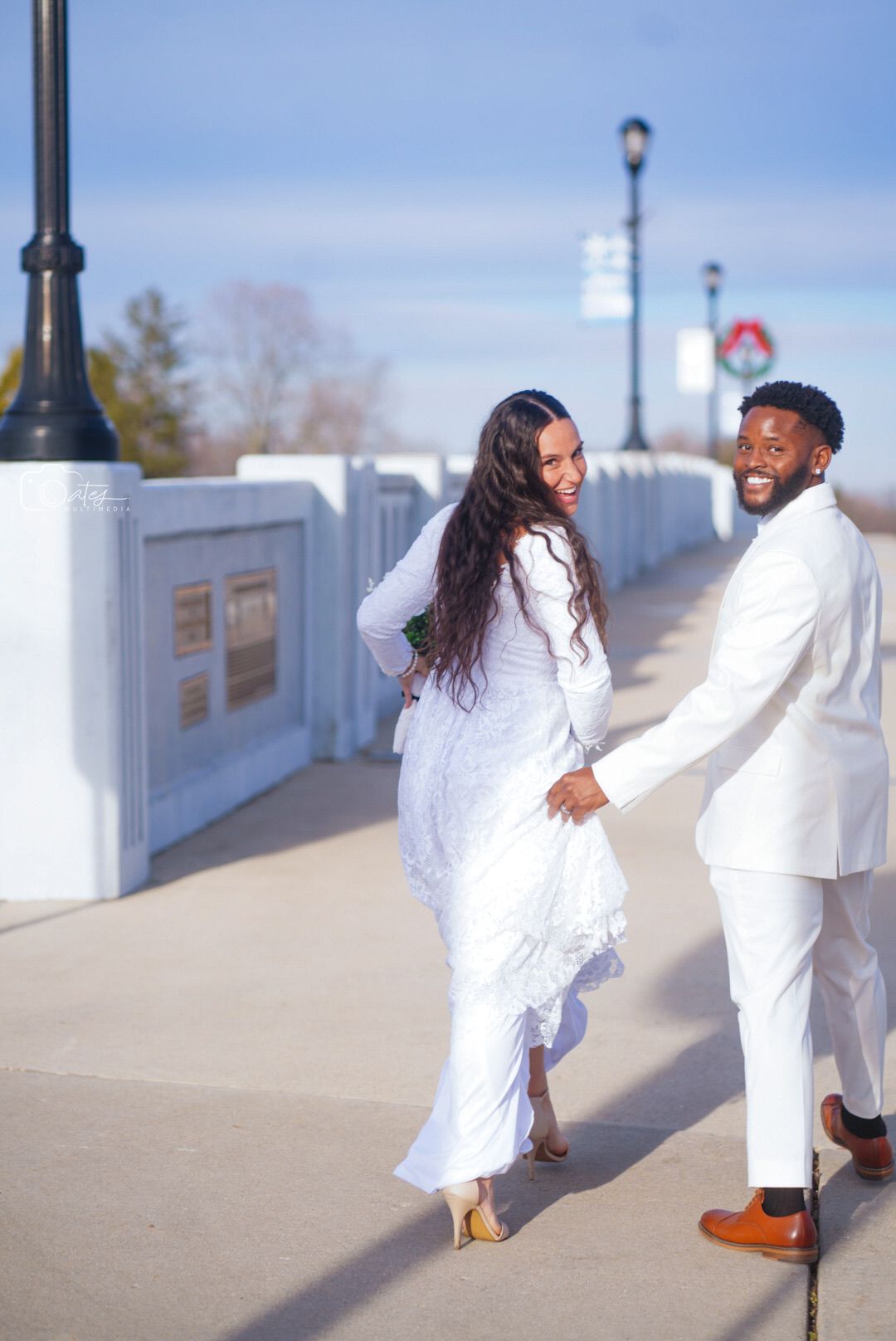Playful wedding couple in all-white outfits dancing on a concrete walkway.