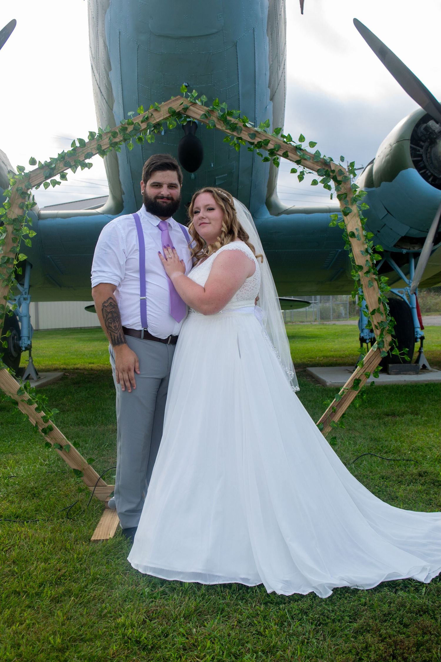 Wedding couple poses by vintage airplane with floral arch decorations on grassy field.