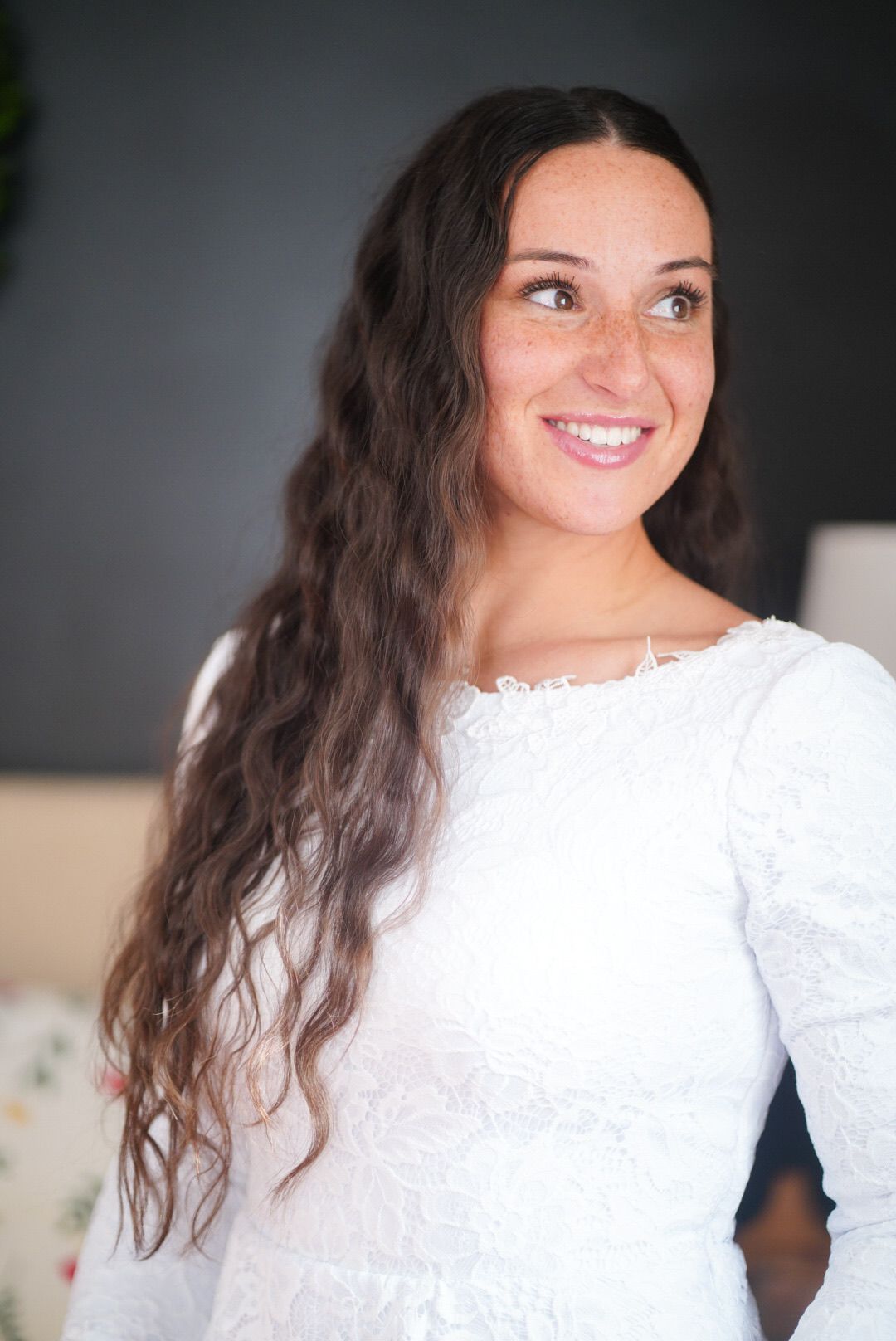 Woman in white lace dress smiles against dark background.