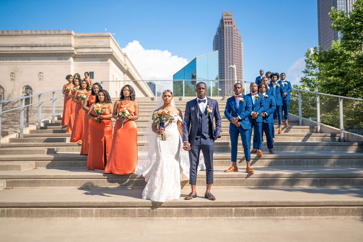 Wedding party lined up on stone steps with bridesmaids in orange and groomsmen in blue suits.