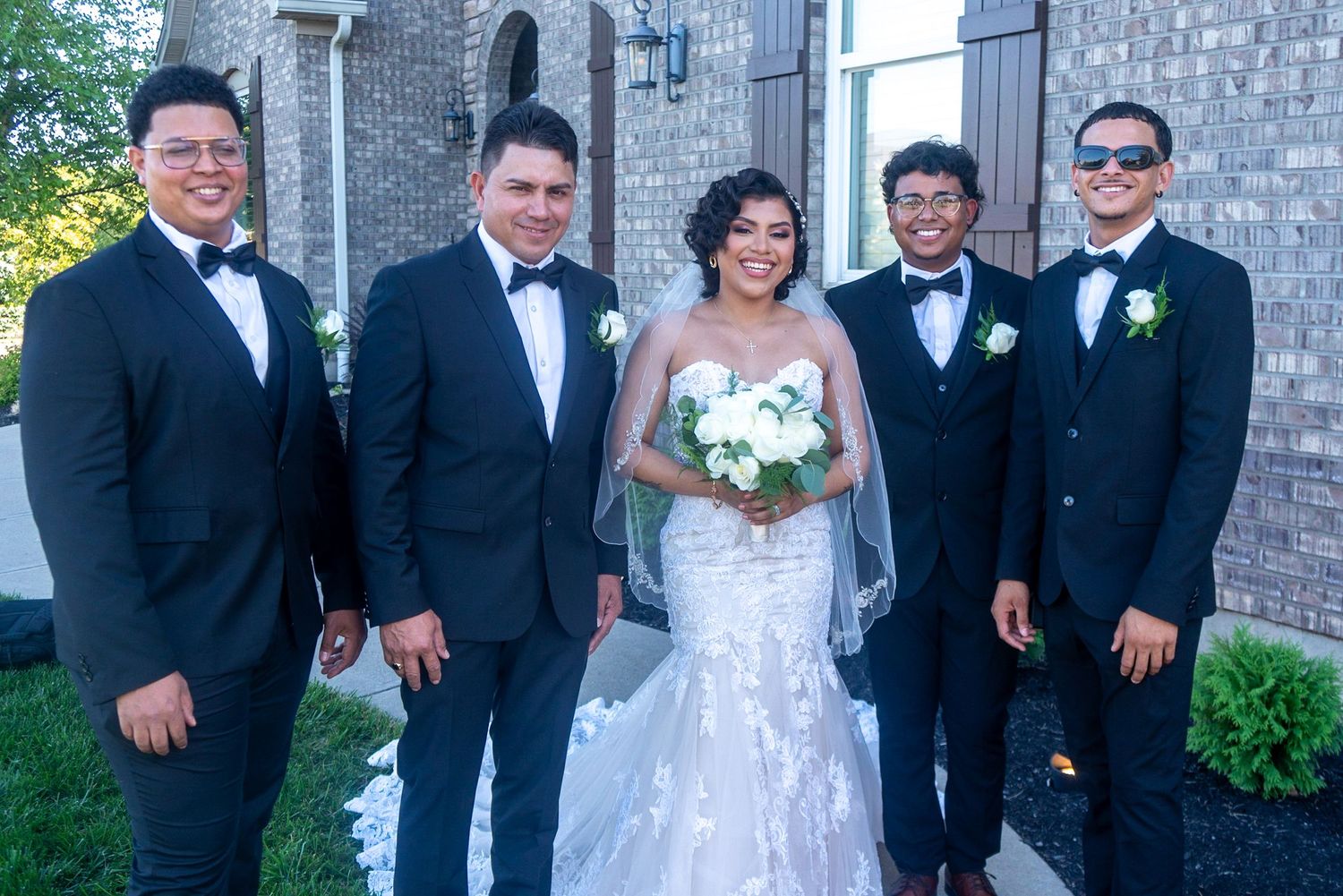 Wedding party members in formal attire pose together against a brick building backdrop.