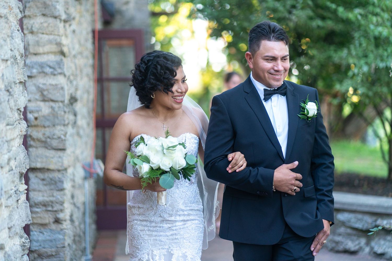 A bride and groom share a romantic moment outdoors near a stone wall during their wedding photoshoot.