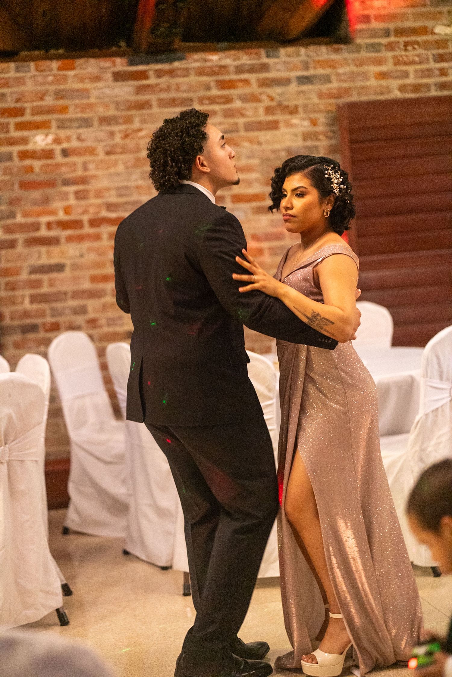 Couple shares a dance moment in a reception hall with white-covered chairs and brick walls.