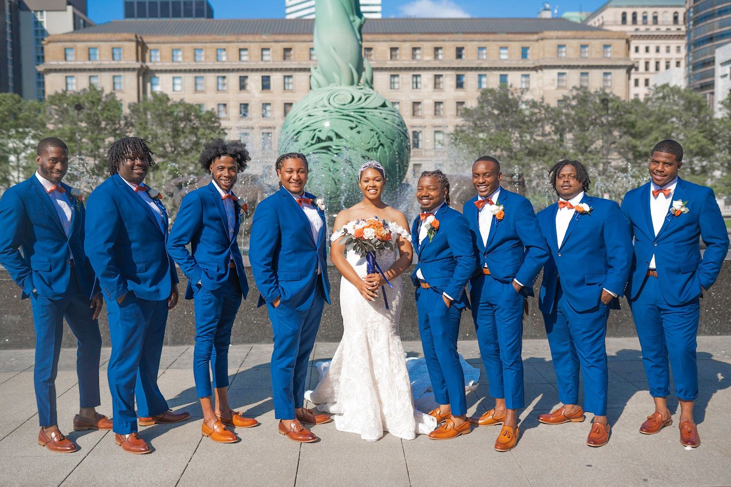 Wedding party in coordinated royal blue suits poses outdoors with city backdrop.
