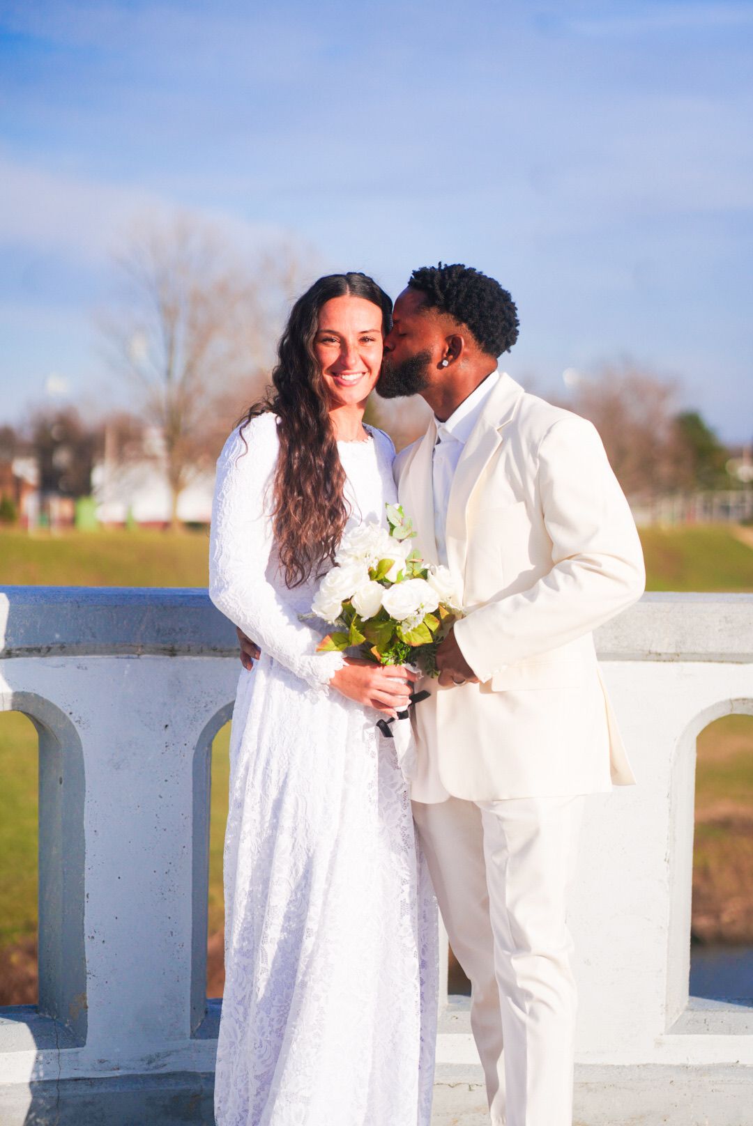 Newlyweds in white attire share intimate moments by a white railing overlooking water.