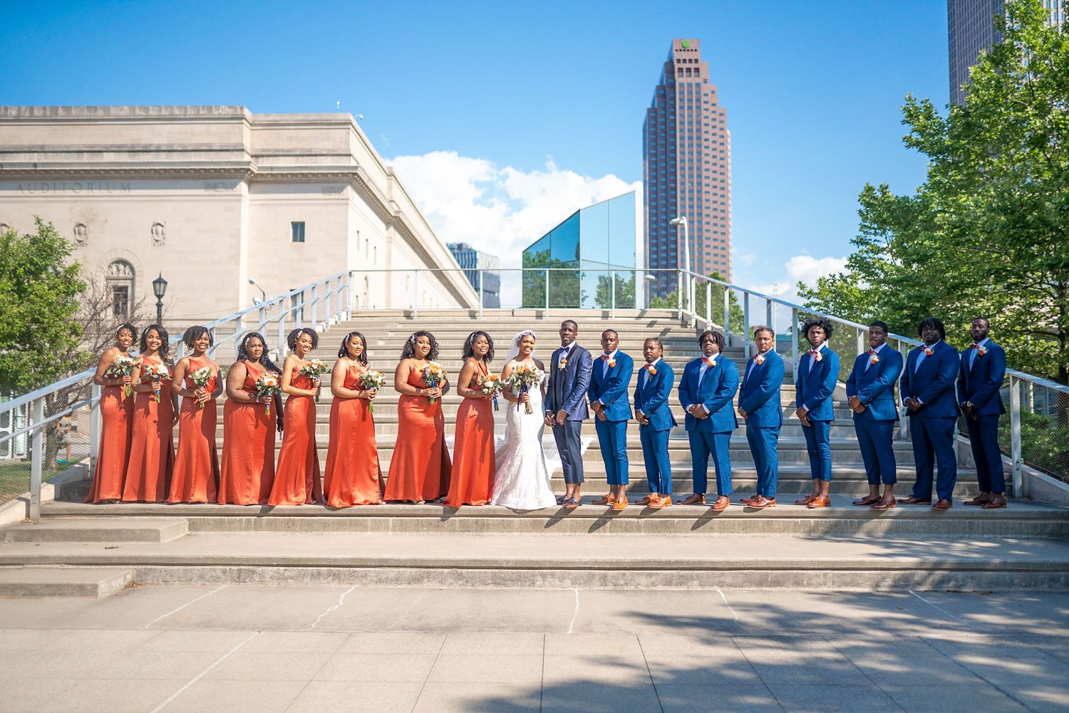 Wedding party in orange and blue attire pose on grand stone steps with city skyline backdrop.