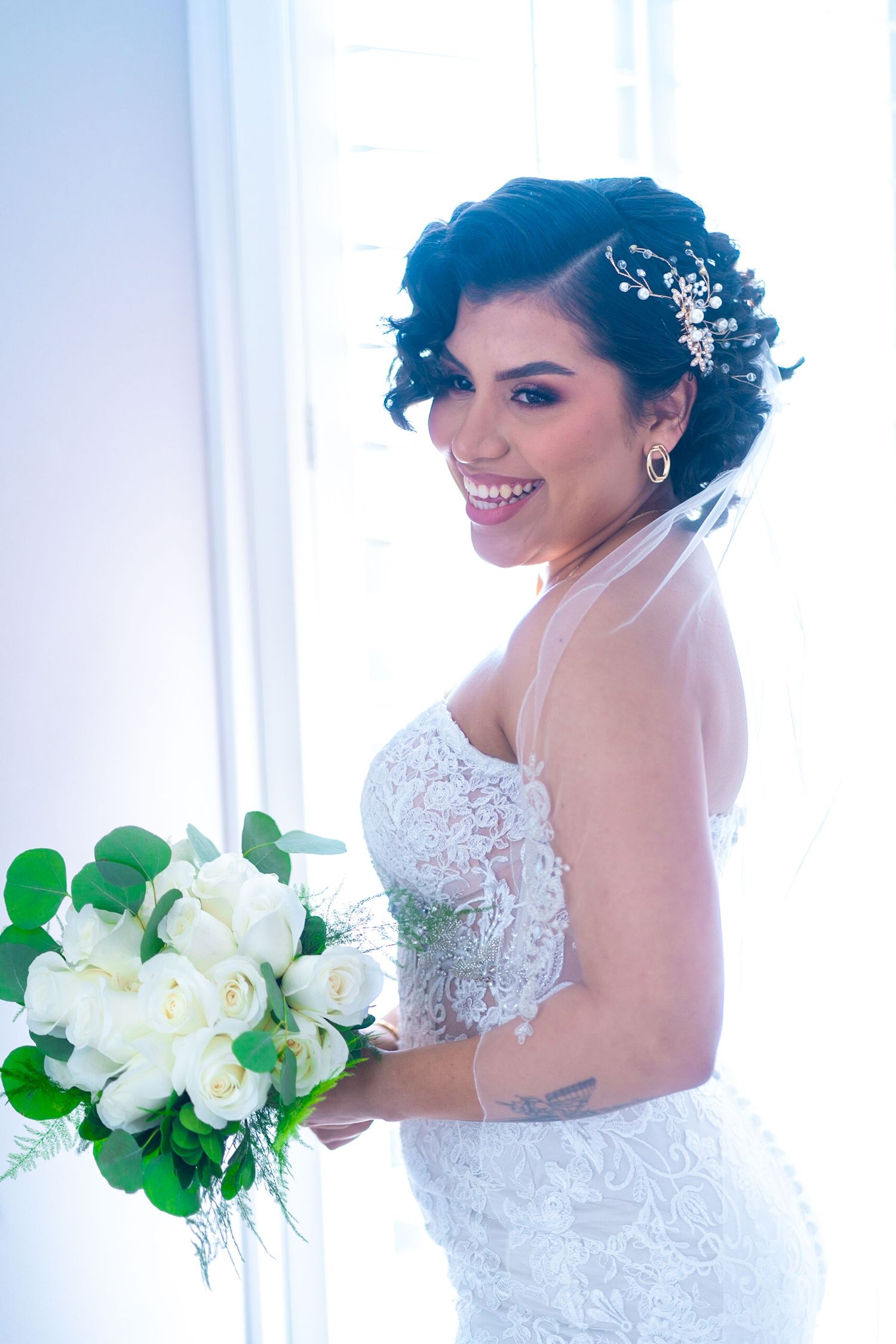 Bride in lace wedding dress holds white floral bouquet against bright window backdrop.