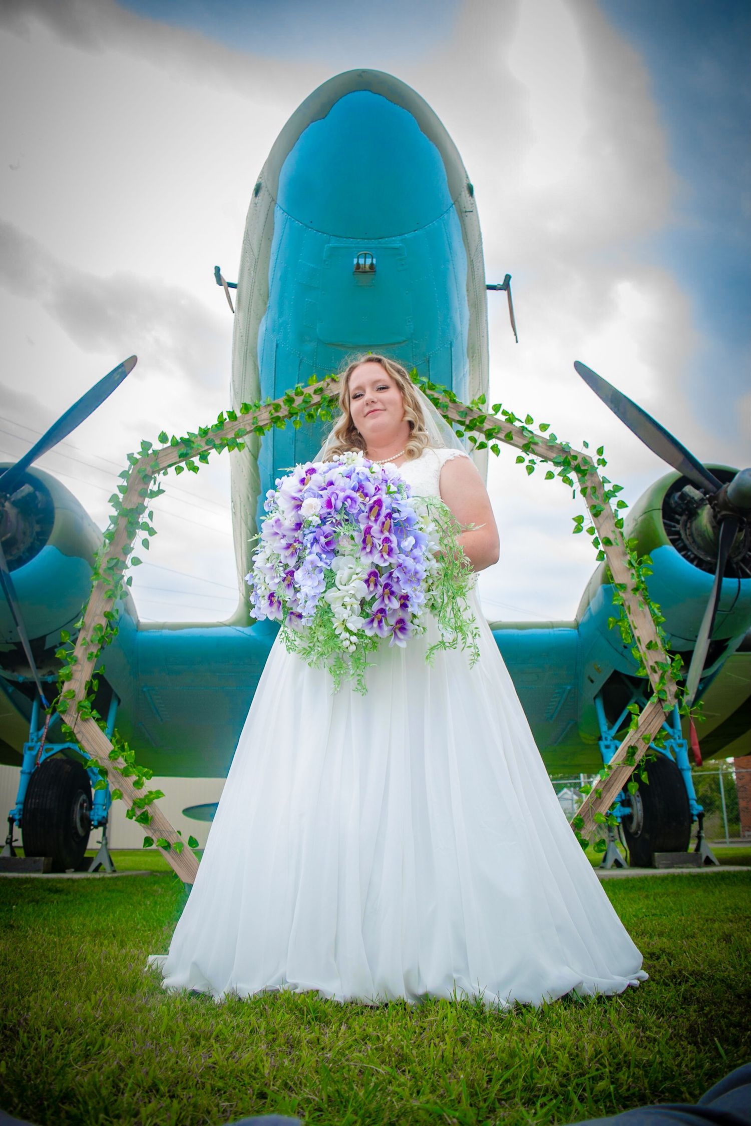 Bridal portrait with vintage airplane and purple floral bouquet in outdoor setting.