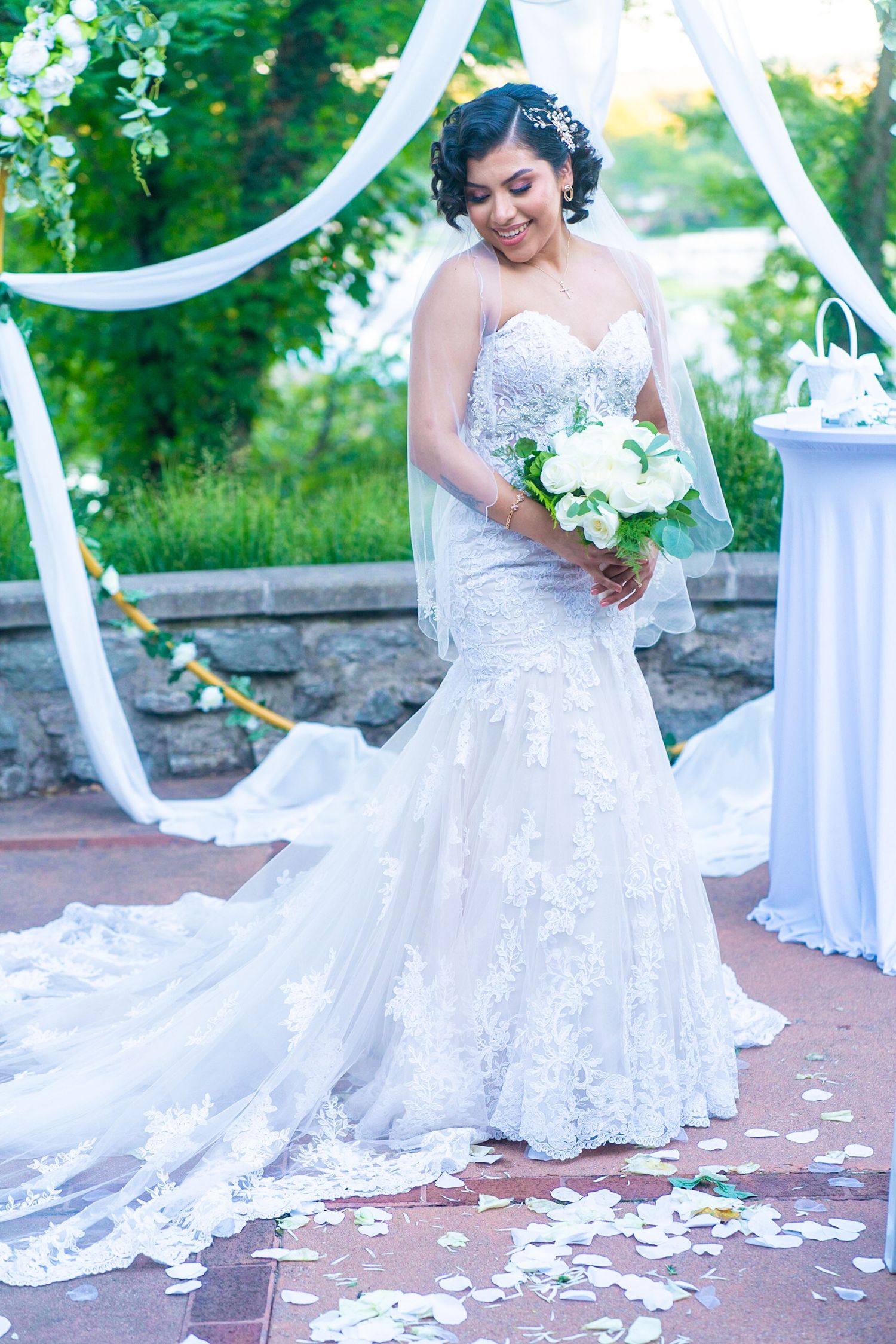 Bride in a flowing lace gown poses under white fabric draping during an outdoor wedding.