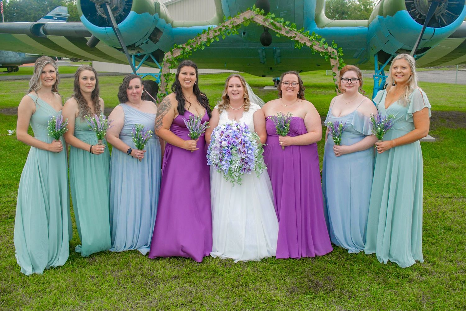 Wedding party in purple and blue dresses poses together in front of vintage airplane.