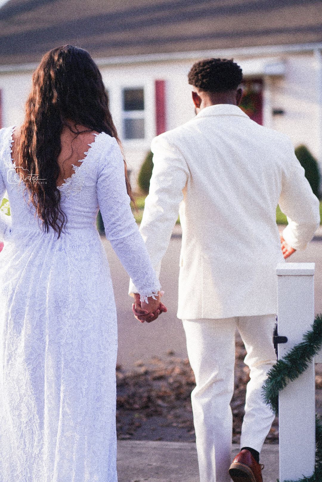 Couple in white wedding attire holding hands while walking along a decorated fence.