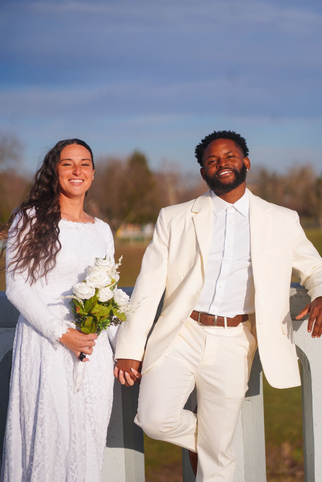A wedding couple poses outdoors in white attire against a natural background.