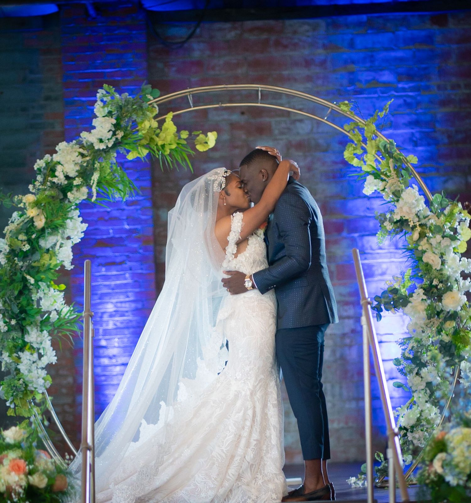 Wedding couple embraces under floral arch with blue uplighting and white flowers.