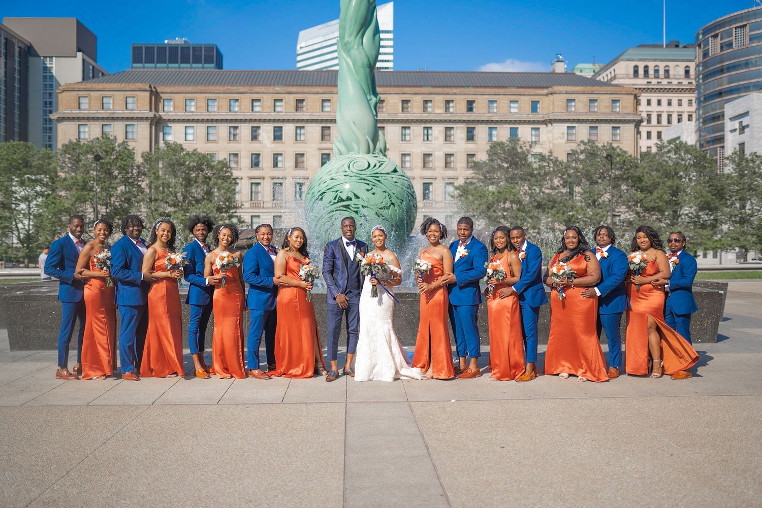 Wedding party in orange bridesmaid dresses and navy suits pose in front of historic building with Statue of Liberty.