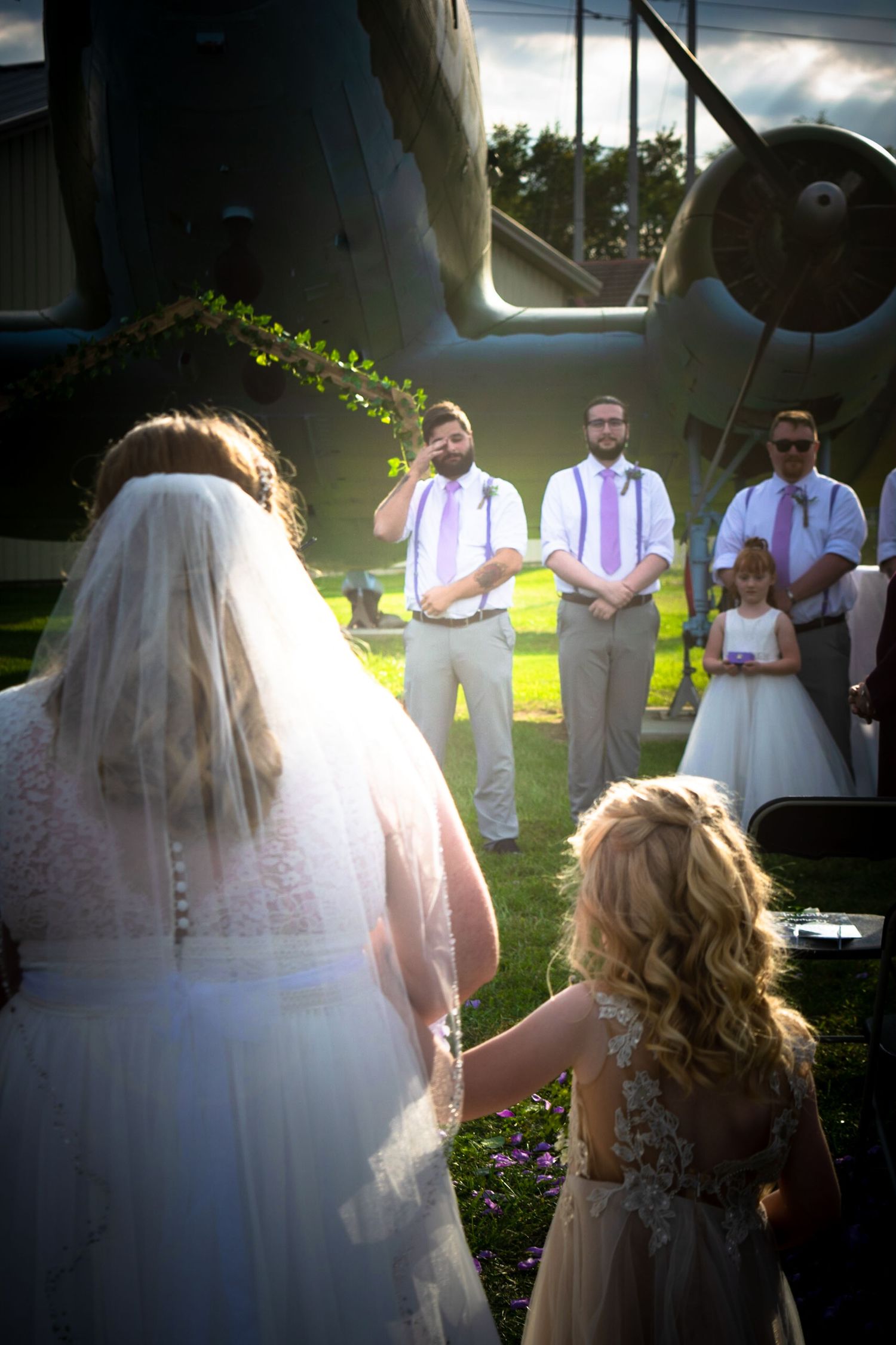 Wedding party gathers for outdoor photos with vintage airplane and natural lighting.