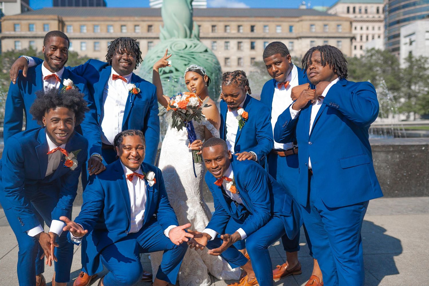 Groomsmen in bright blue suits pose playfully by fountain in urban setting.