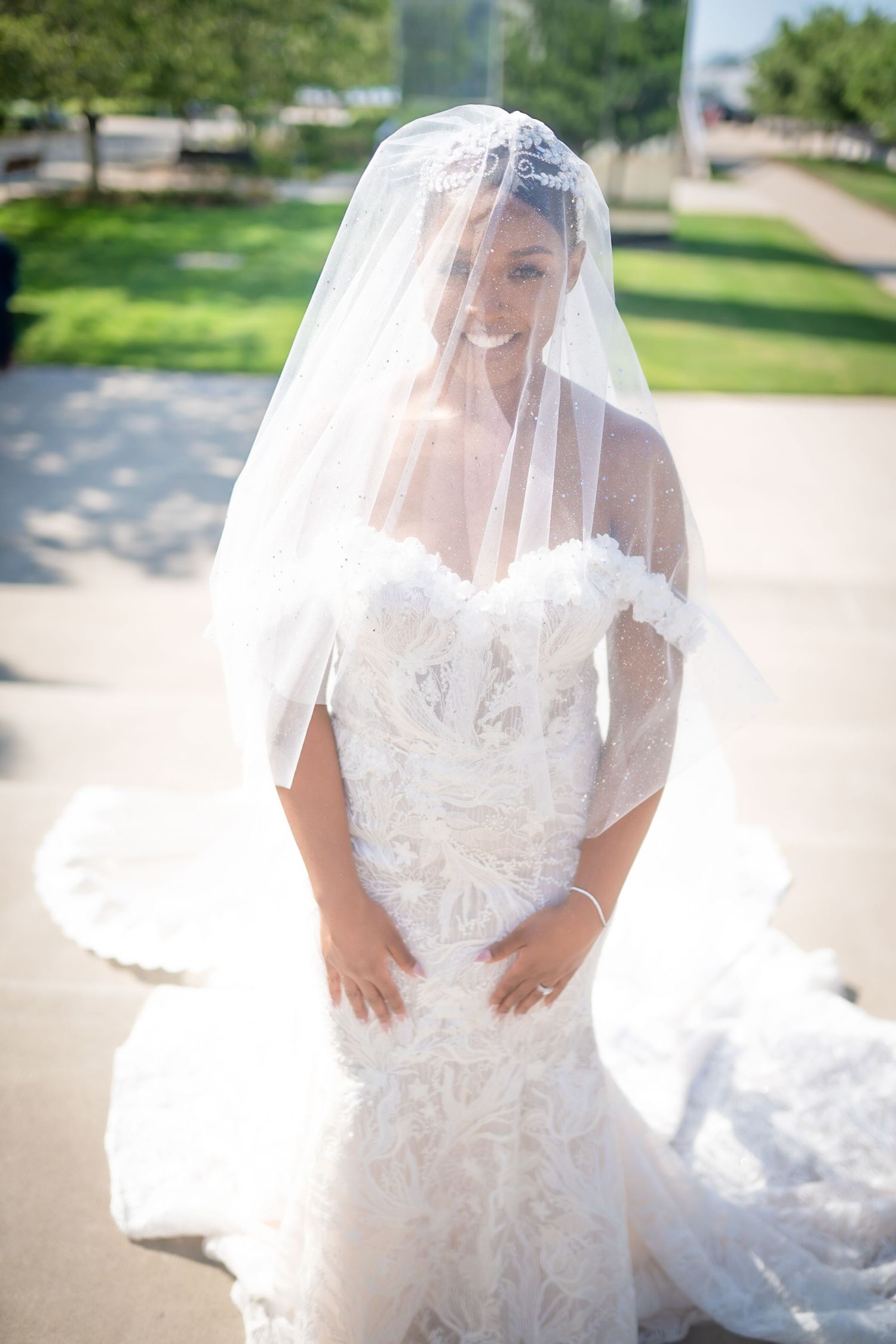 White lace wedding dress with flowing veil captured in soft sunlit outdoor setting.