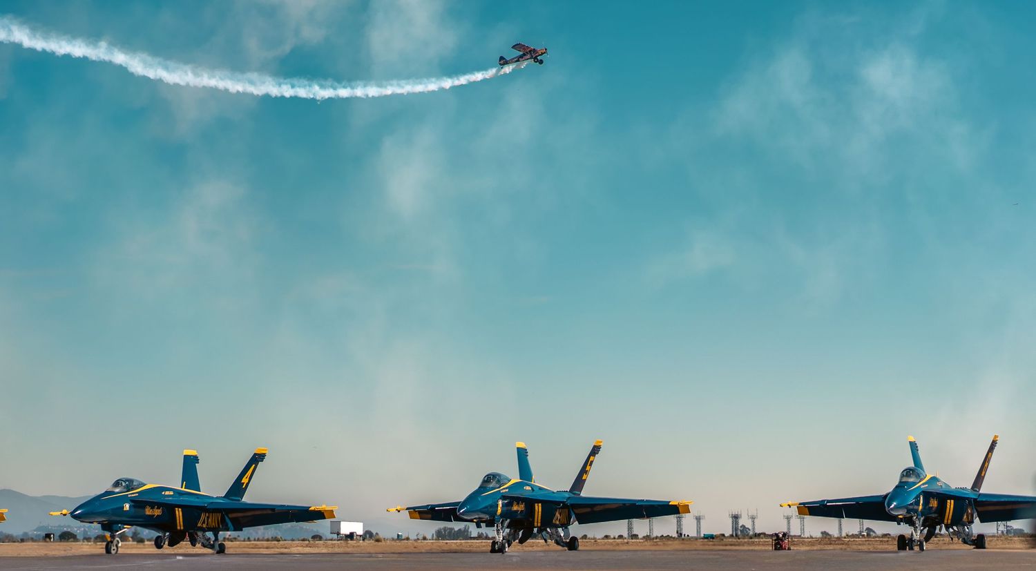 Blue Angels jets on runway with aerobatic plane performing overhead against bright blue sky.