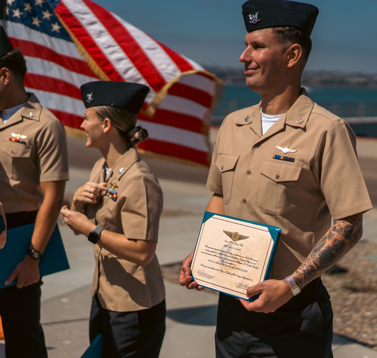 Military personnel in dress uniforms at ceremony with American flag in background.