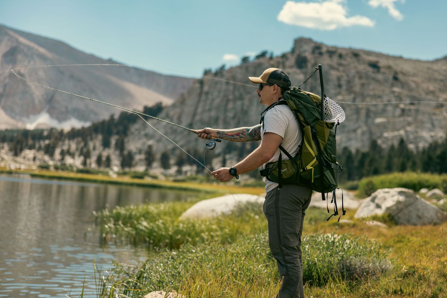 Person with backpack fly fishing in mountain lake surrounded by scenic landscape.