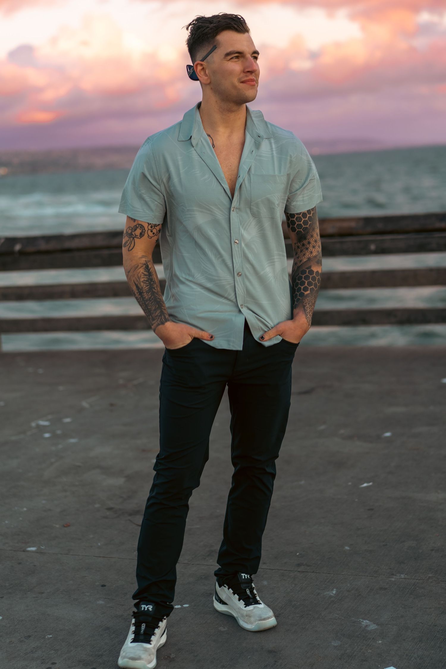 Person in mint button-down shirt and dark jeans poses on beach at sunset with tattoo-covered arms.