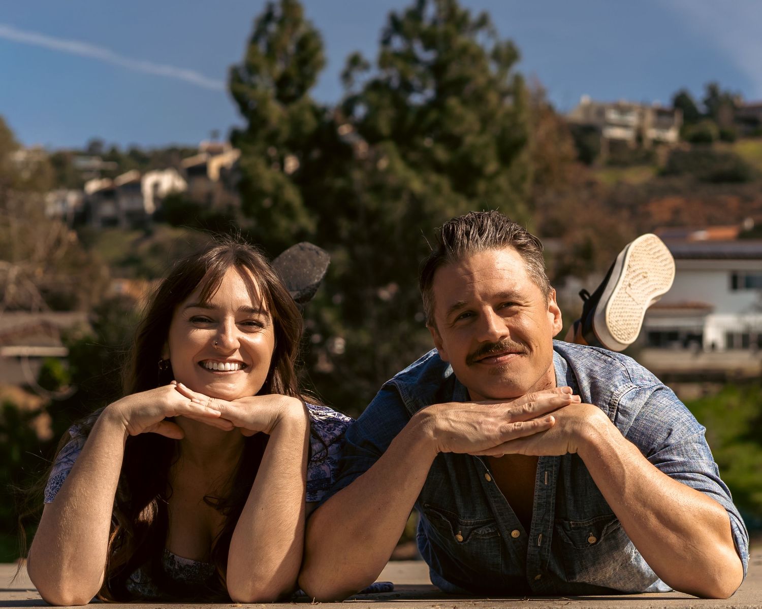 Two people lying down and smiling while looking at the camera outdoors with mountains in the background.