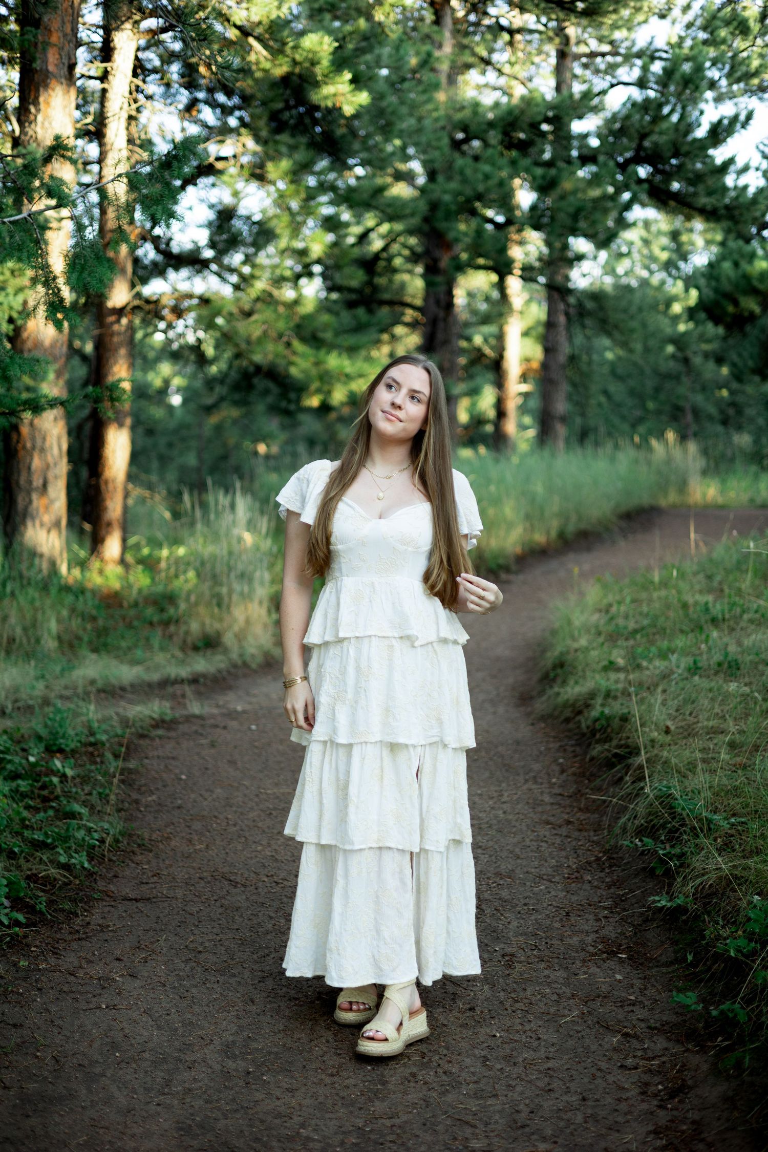 A person in a flowing white dress stands on a dirt path surrounded by tall pine trees in a forest setting.