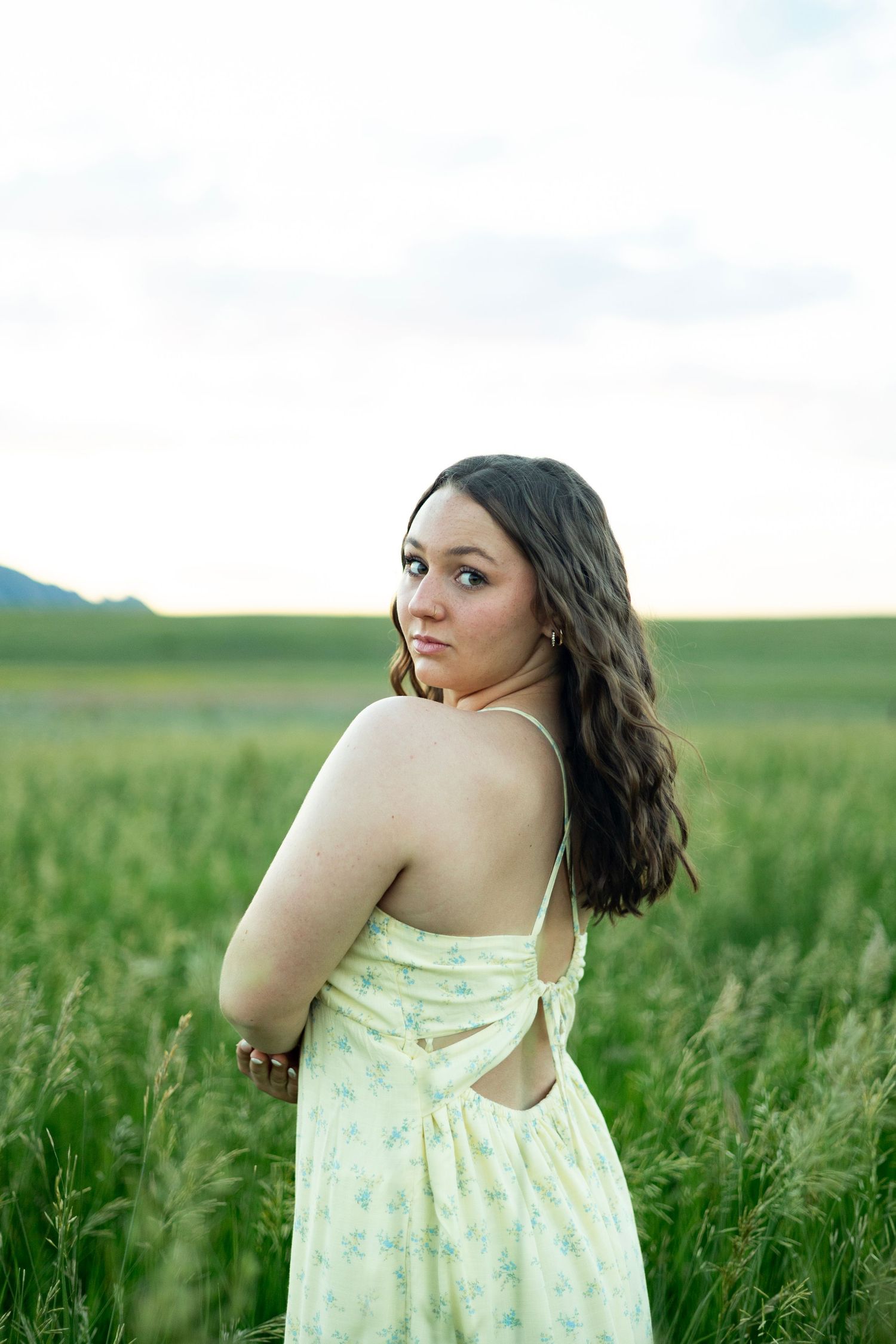 Person in yellow floral dress poses thoughtfully in a lush green field on a summer day.