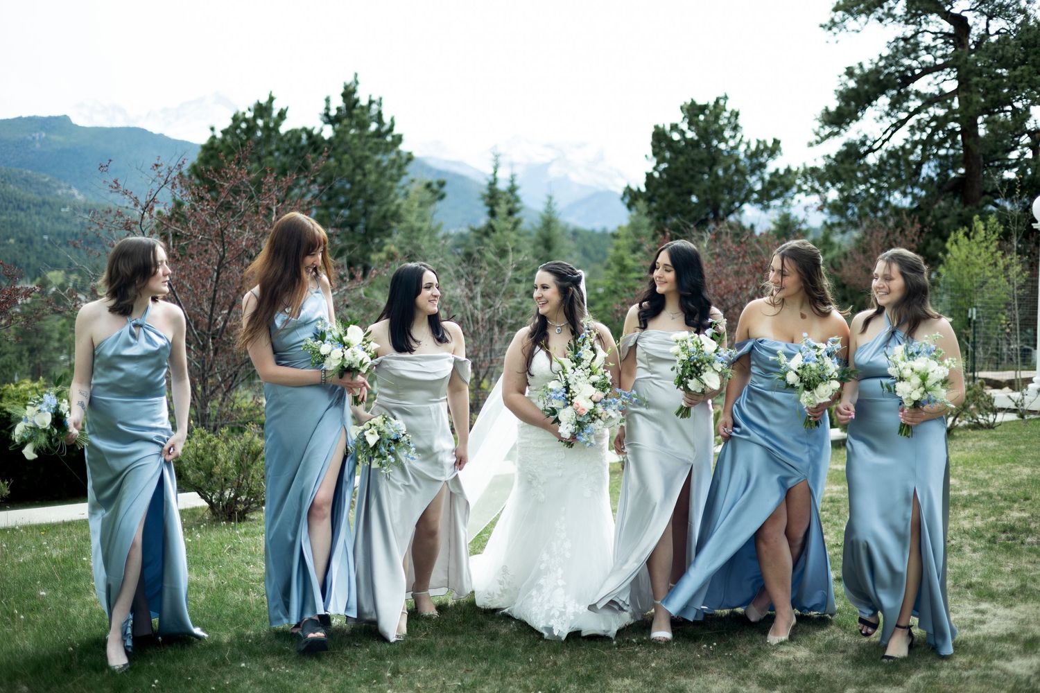 Bridesmaids in flowing light blue dresses hold bouquets while posing on a grassy hill with mountain views in background.