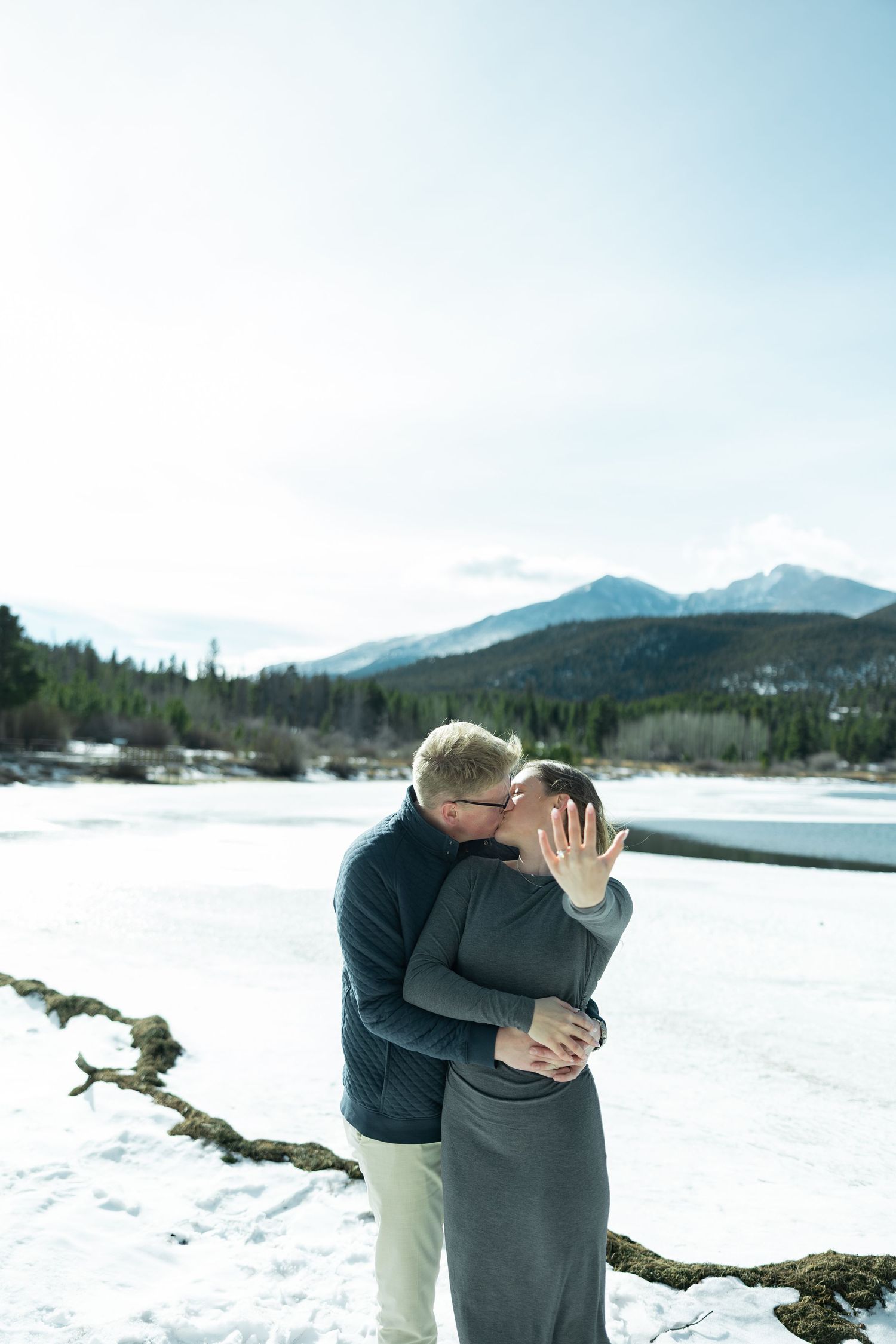 Two people embrace in a romantic moment against a snowy winter mountain landscape with evergreen trees.