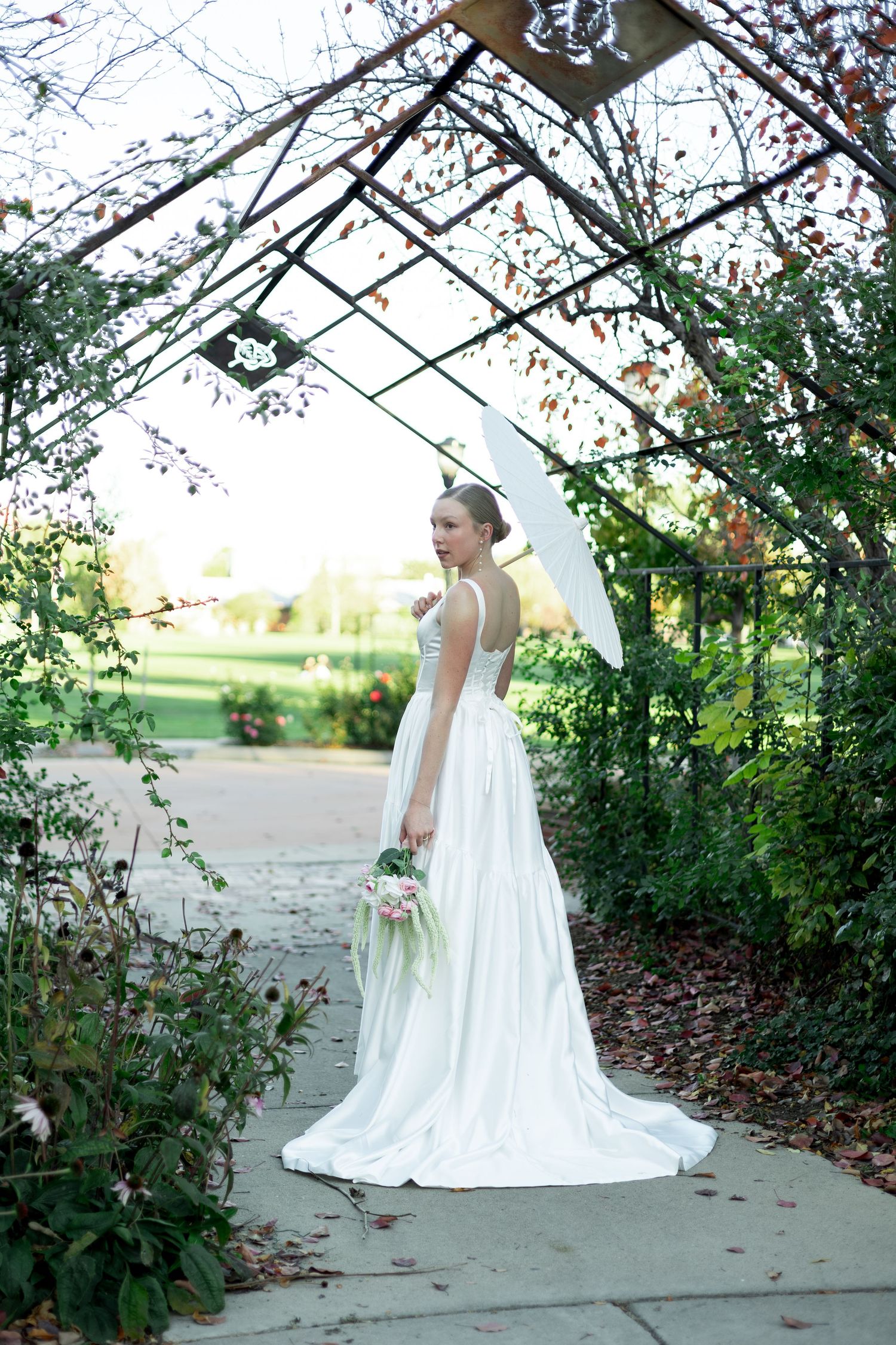 Bride in white gown holding bouquet stands under garden arbor with lush greenery on stone pathway.