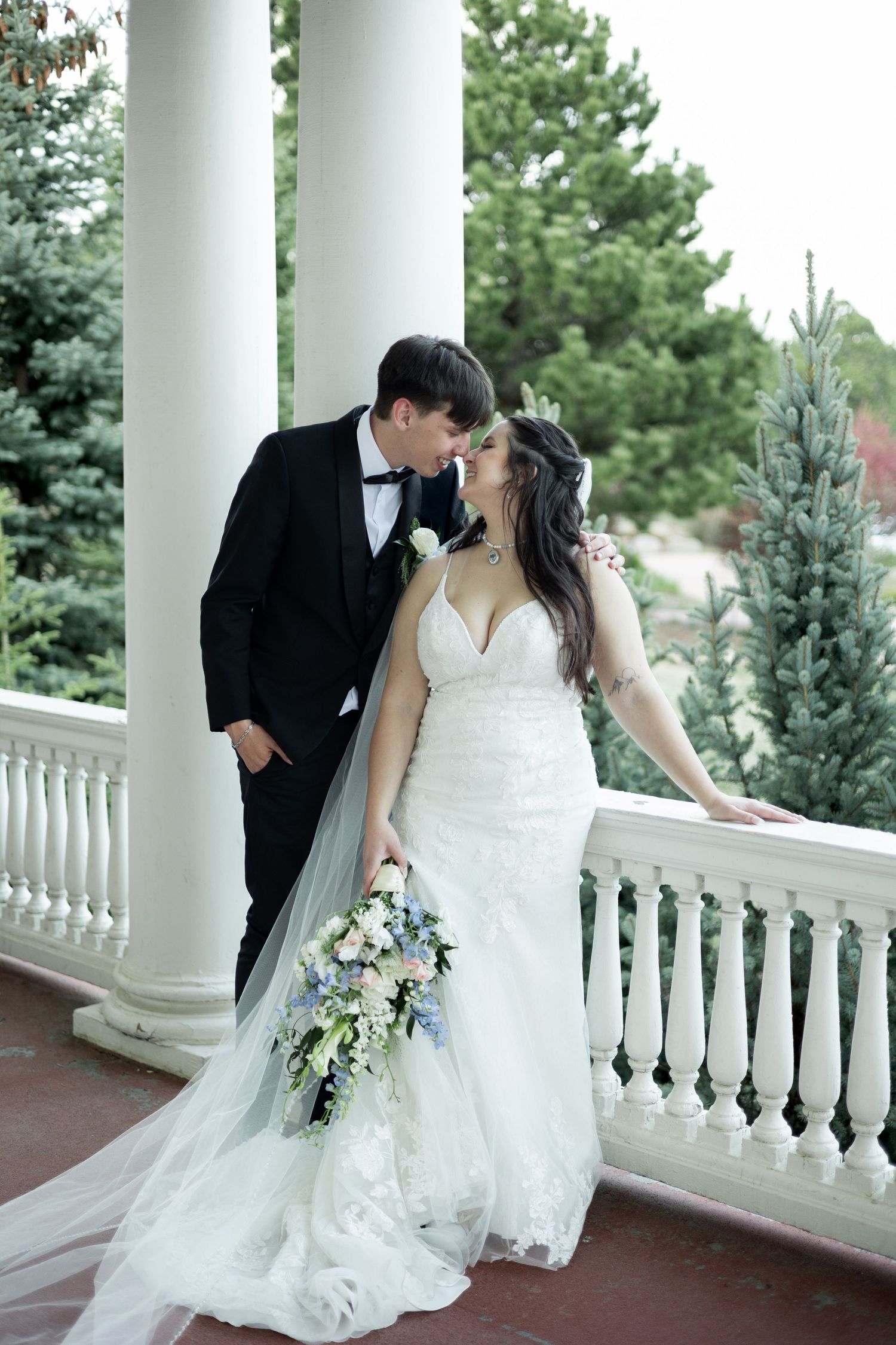 Romantic wedding couple embraces on an elegant white columned porch with pine trees in background.