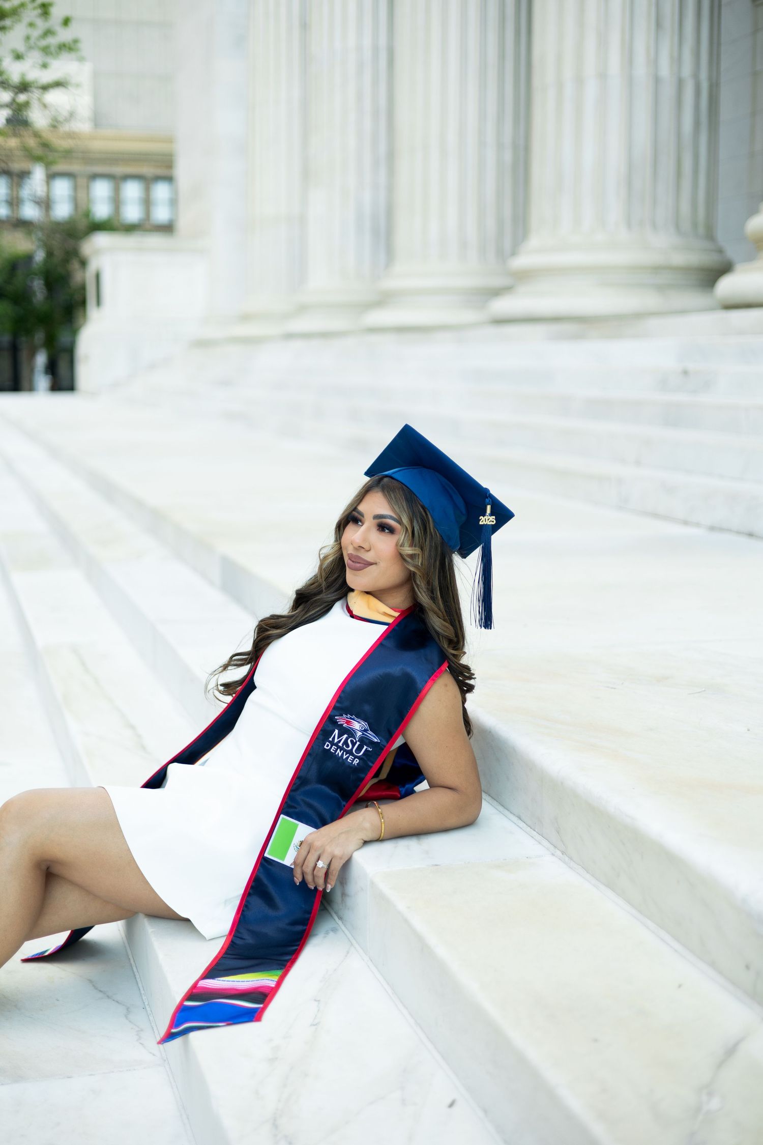 Graduate in blue cap and gown with graduation sash sits on white marble steps for portrait photo session.