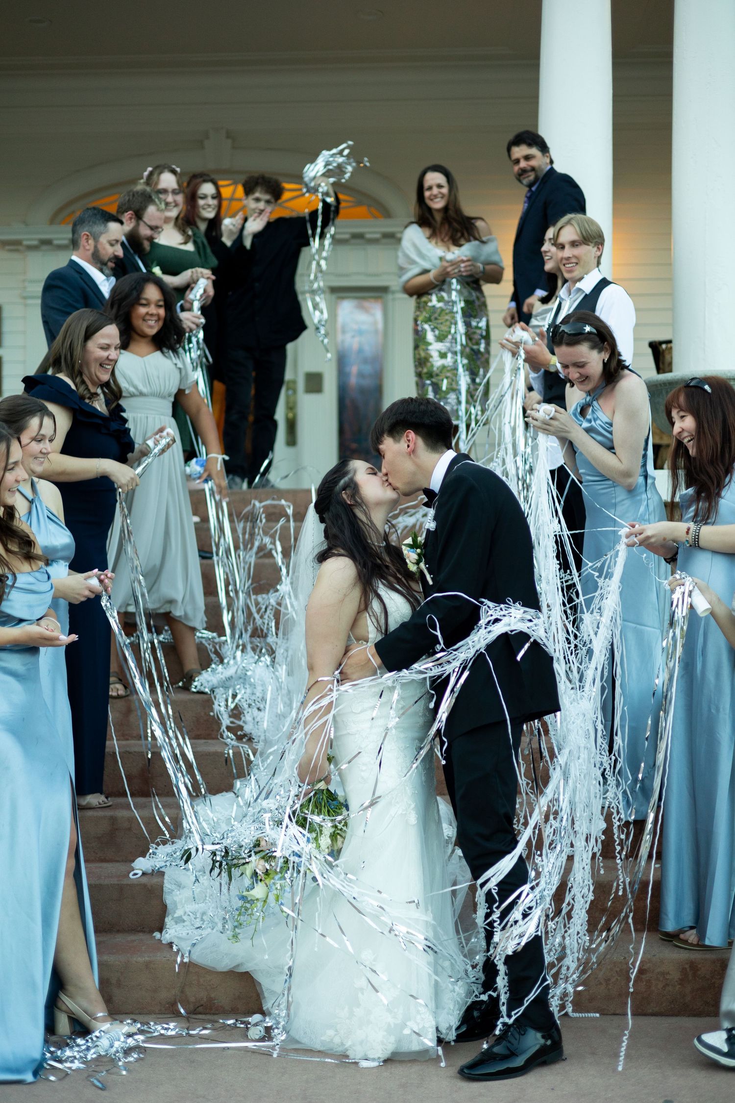 A kissing couple at their wedding celebration gets showered with silver streamers as wedding party members look on and cheer.