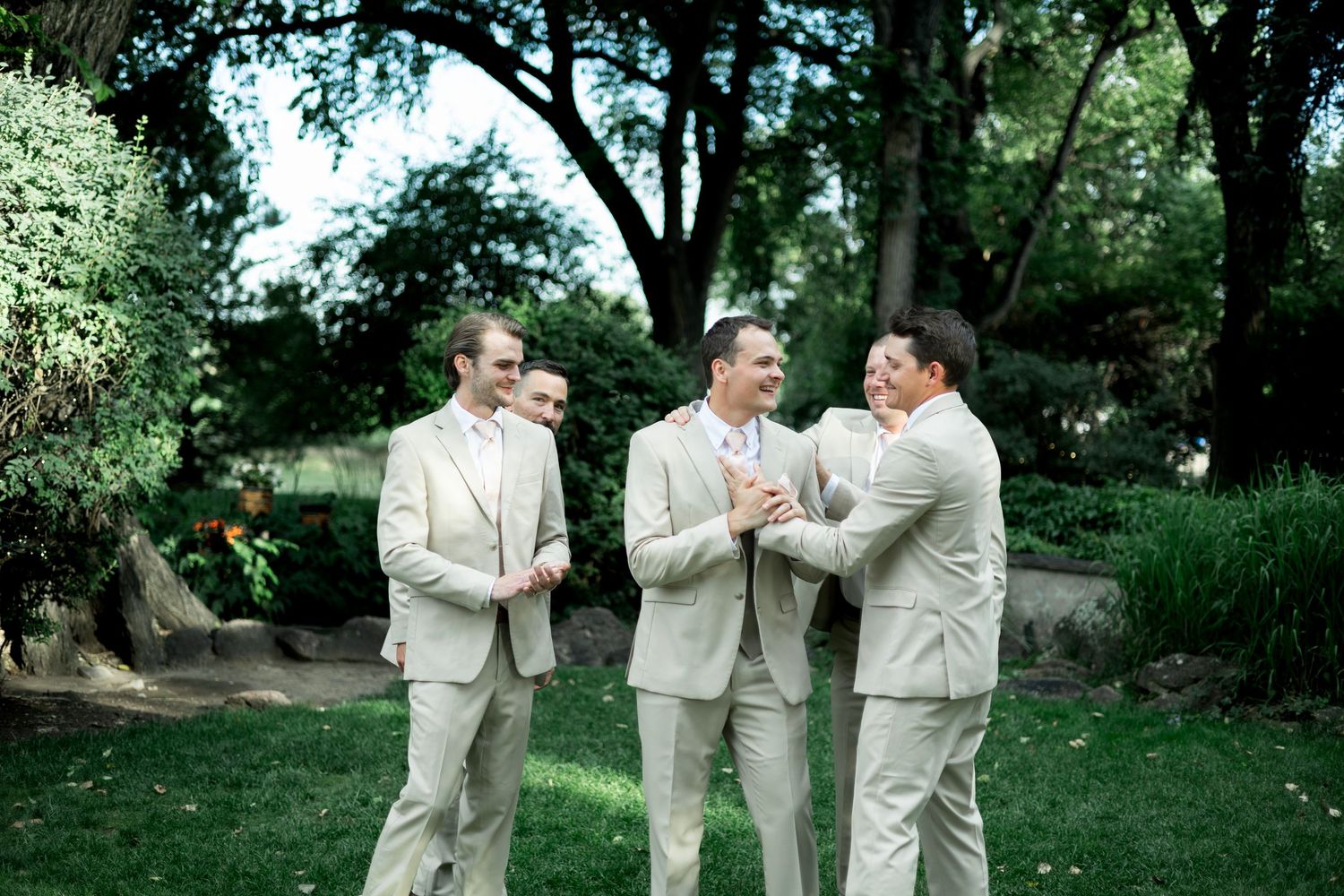 Groomsmen in light beige suits gather outdoors under trees for wedding day photos.