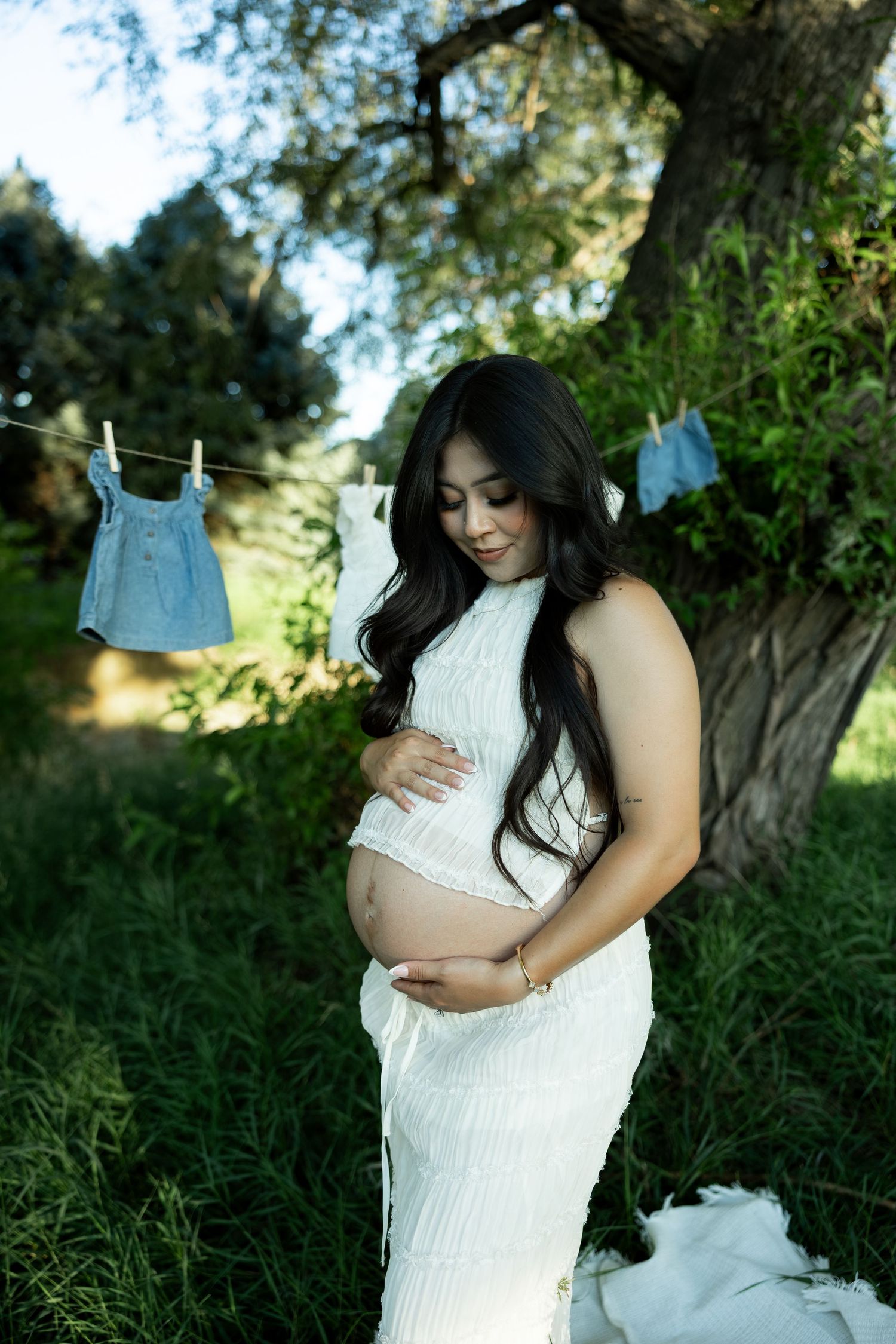 Pregnant woman in white dress cradling baby bump outdoors, with baby clothes hanging on a clothesline behind her.