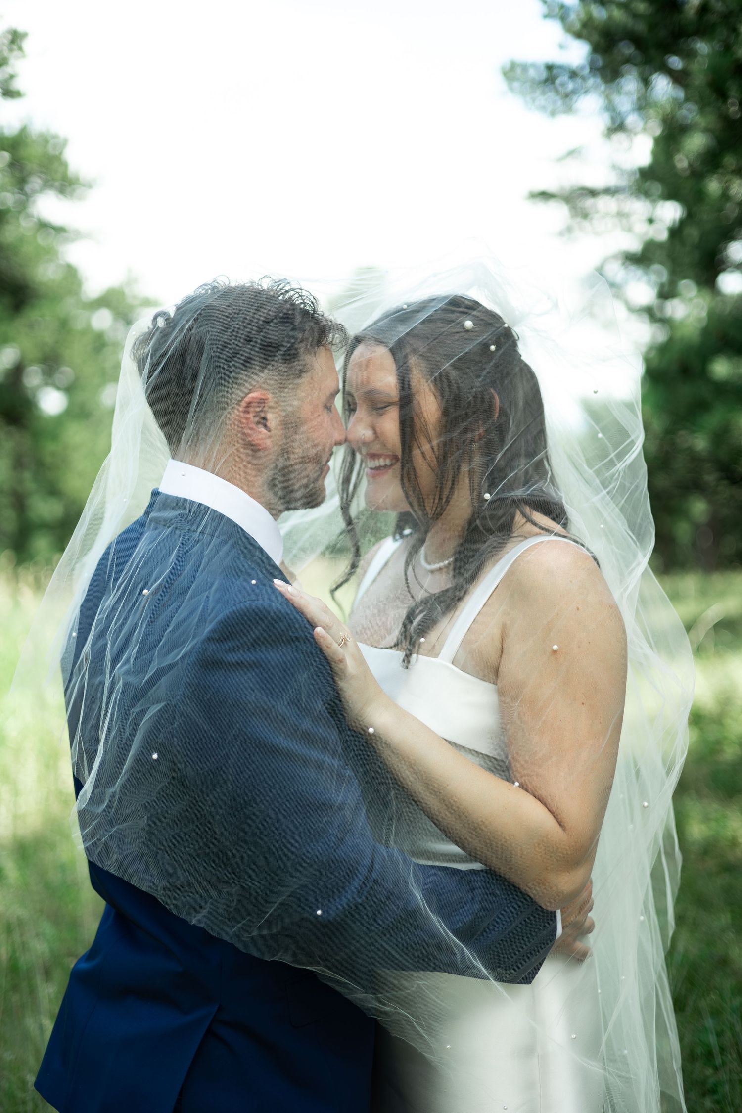 Bride and groom share a romantic moment under a wedding veil outdoors, surrounded by lush greenery.
