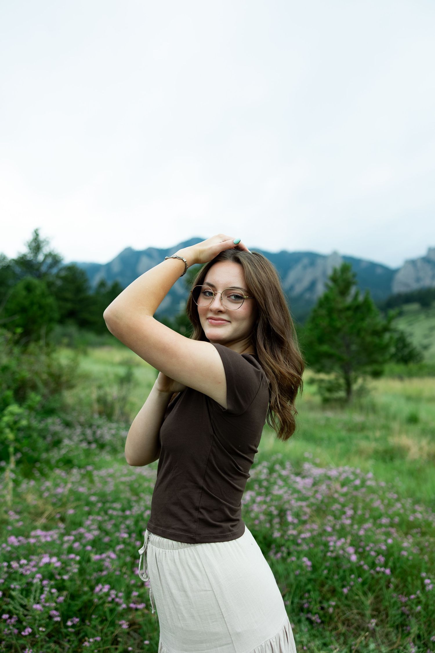 Young woman in glasses and brown top poses in a wildflower meadow with mountain backdrop.