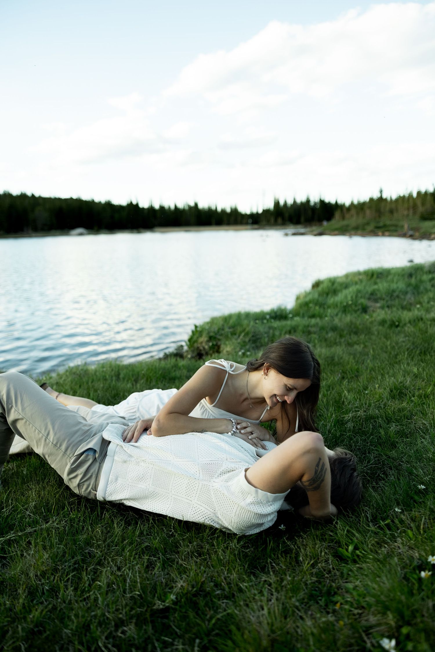 Romantic couple lying on green grass by a serene lake, woman leaning over man in tender embrace outdoors.