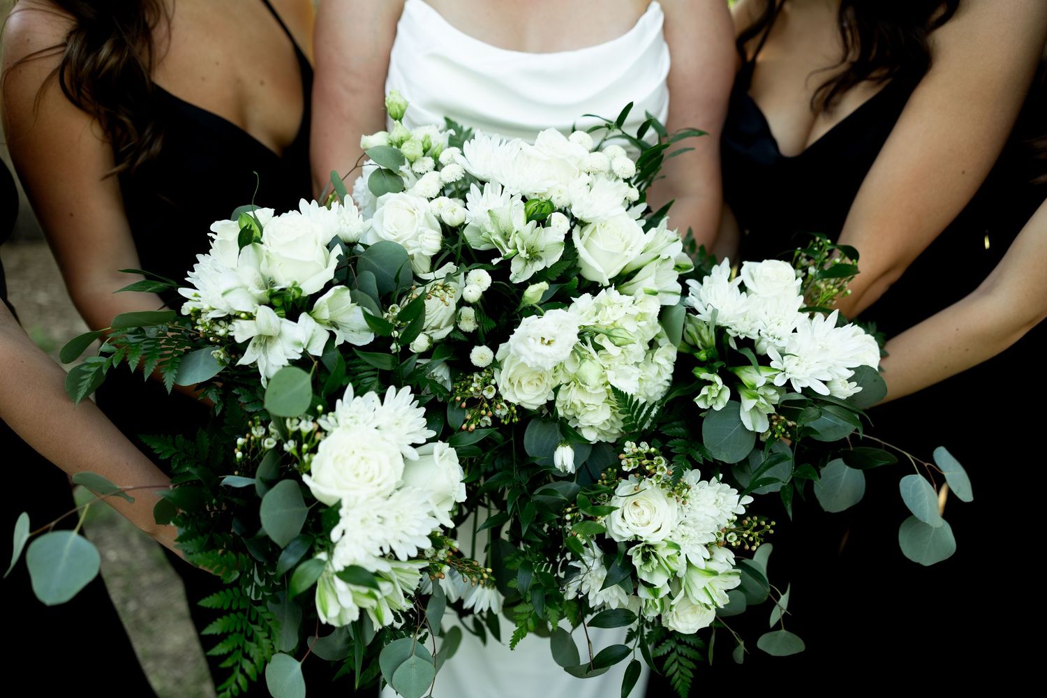 Bride holding a lush white floral bouquet with eucalyptus, flanked by bridesmaids in black dresses.