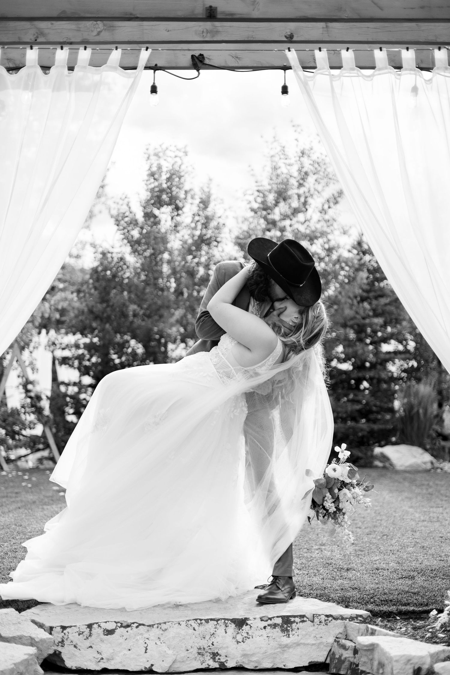 Black and white photo of bride and groom sharing first kiss under draped wedding altar outdoors.