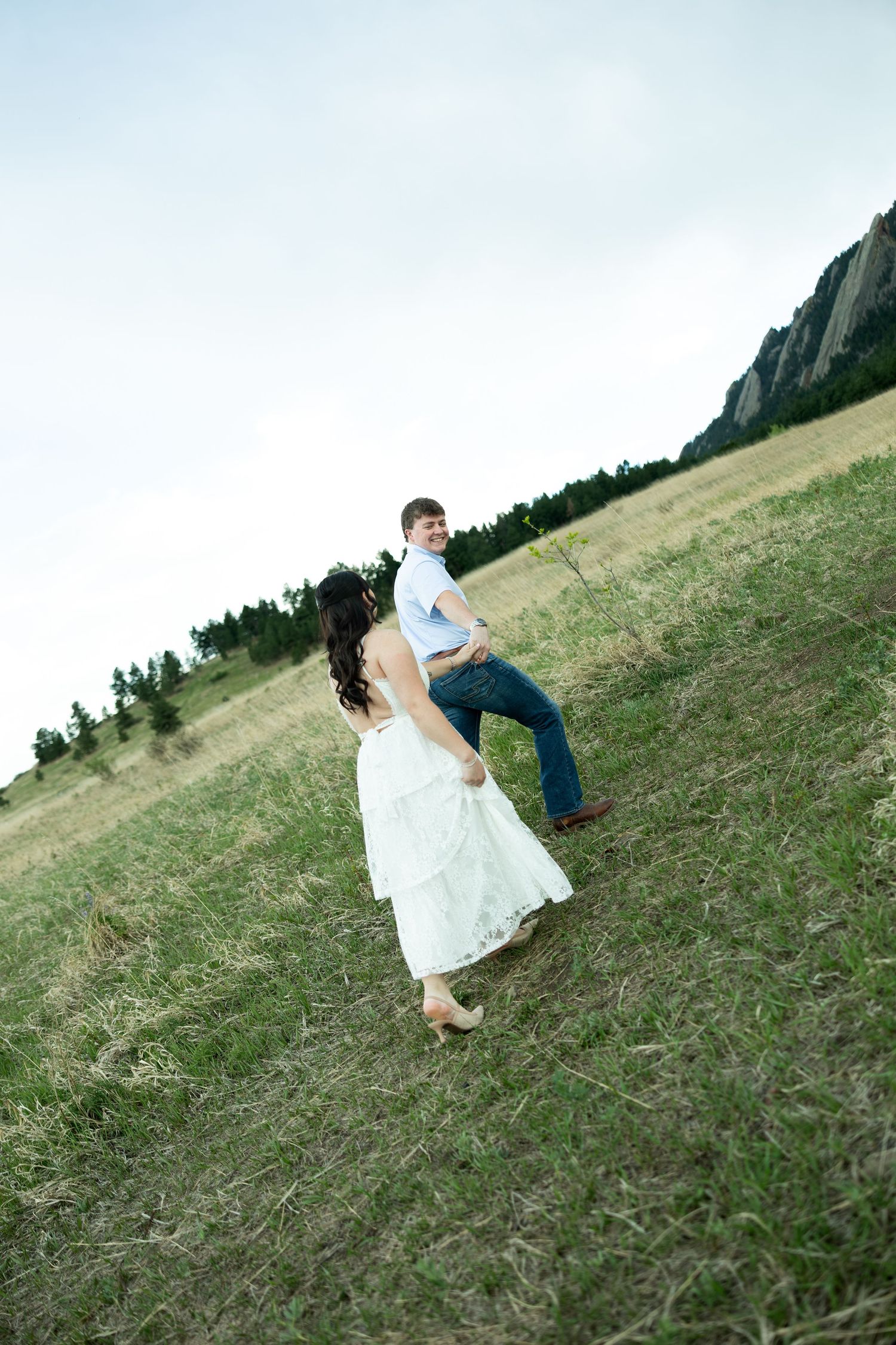 Couple in wedding attire dancing joyfully in a green meadow with rocky mountains and trees in the background.