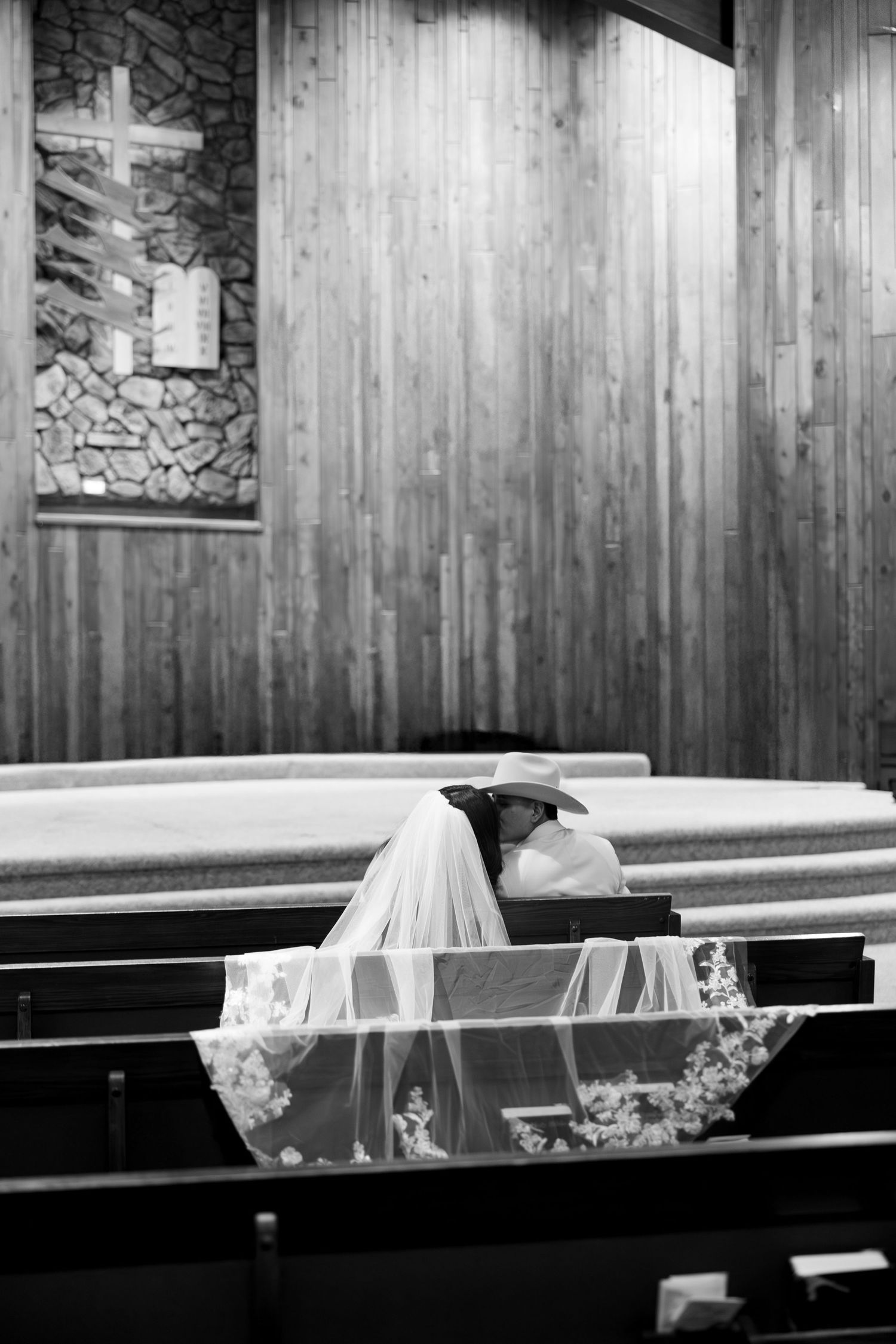 A wedding veil and dress displayed across wooden church pews in black and white.