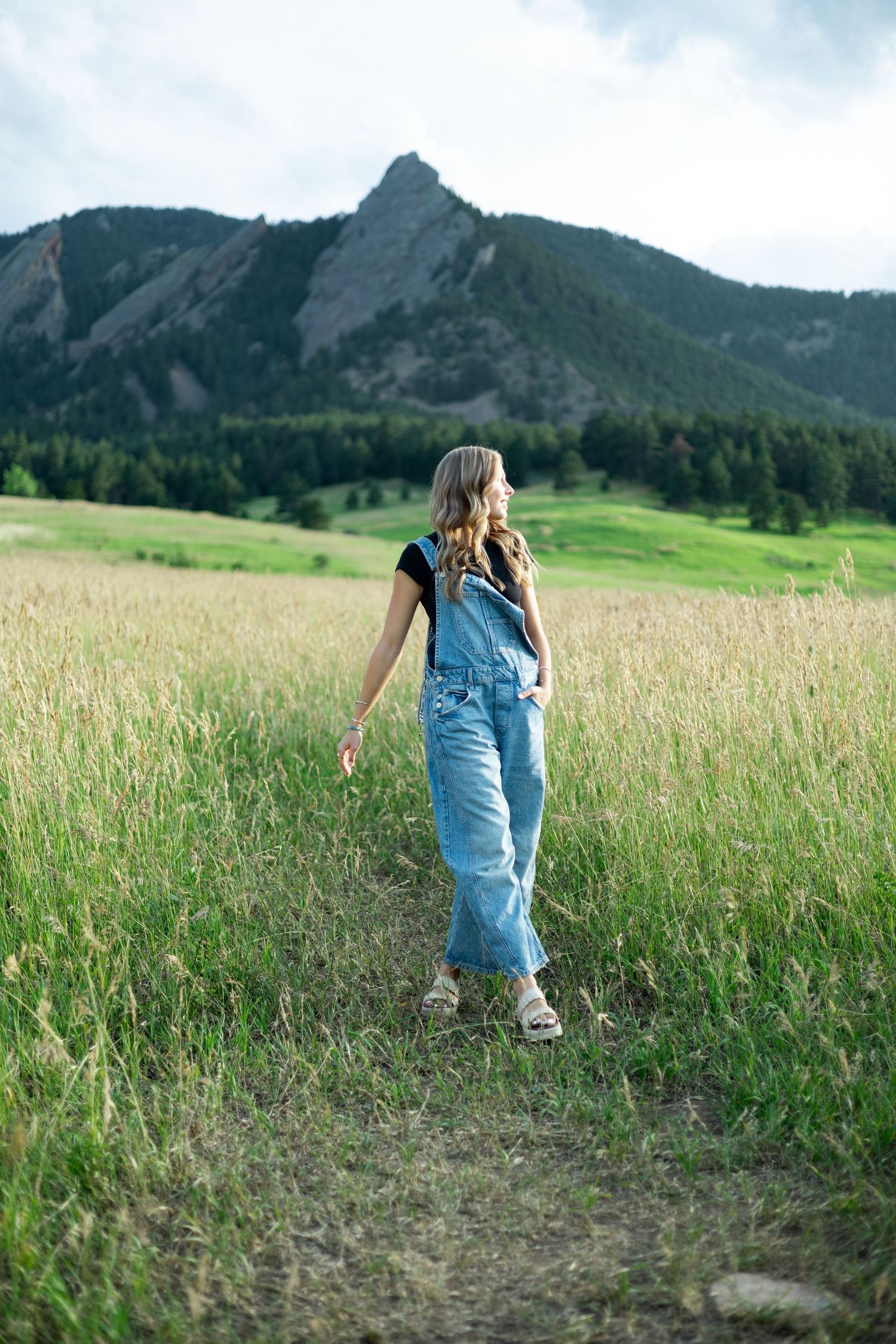 A young woman in denim overalls walks through a green meadow with rocky mountain peaks and pine trees in the background.